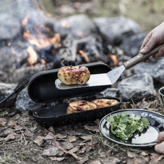 Flexible spatula with a polished metal blade and a smooth wooden handle lifting cooked food outdoors by a campfire, designed for Kitchen & Food, Grilling, and Cooking Helpers collections.