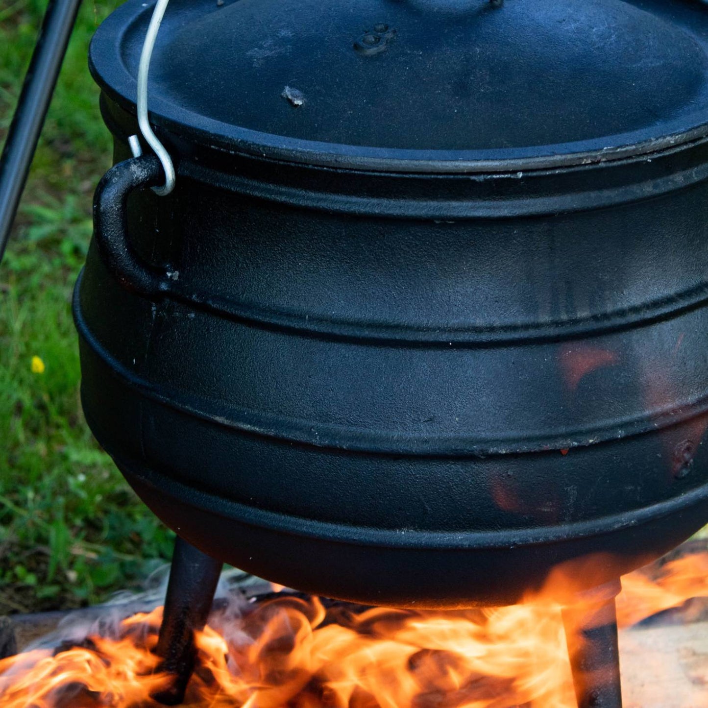 Handcrafted cast iron campfire kettle with a rounded ribbed body, fitted lid, and tripod legs, hanging over an open flame in an outdoor setting for Farm, Garden & Tool, Camping, Best Sellers, Promo Eligible collections.