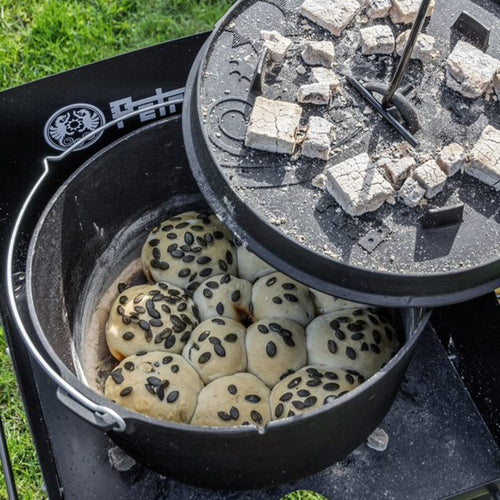 Black cast iron Dutch oven with flat base baking seeded bread rolls over hot charcoal briquettes on its lid outdoors, perfect for Kitchen & Food, Cookware, Cast Iron, Best Sellers, Promo Eligible collections.