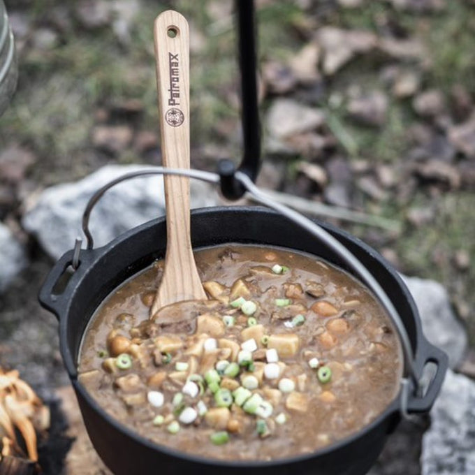 Black cast iron Dutch oven with flat base outdoors filled with hearty stew garnished with chopped green onions, featuring a wooden spatula with a branded handle, in Kitchen & Food, Cookware, Cast Iron, Best Sellers, Promo Eligible collections.