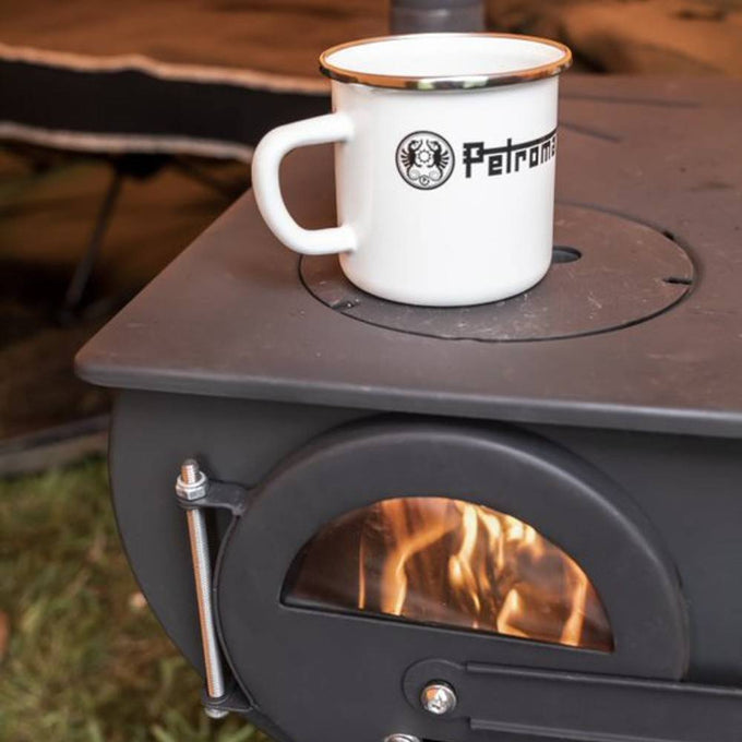 White enamel steel mug with metal rim and black logo, resting atop a wood-burning stove with visible flames, suitable for Farm, Garden & Tool, Camping, Kitchen & Food, Brewing Coffee and Tea collections.