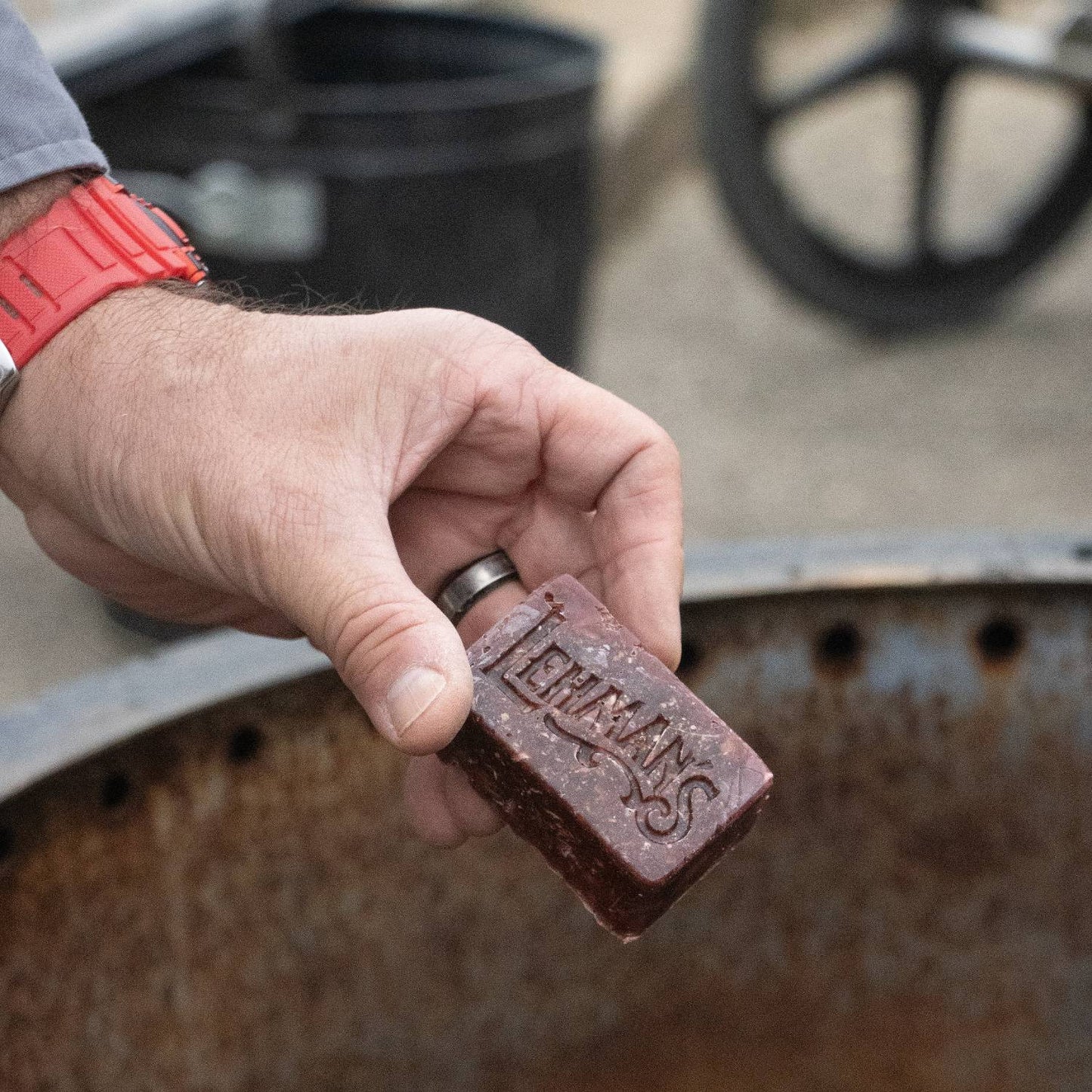 Handcrafted reddish-brown fire starter block embossed with Lehman’s logo, held above a metal fire ring, designed for easy, non-toxic fire lighting in stoves, camping, and hearth accessories from USA Made collection.