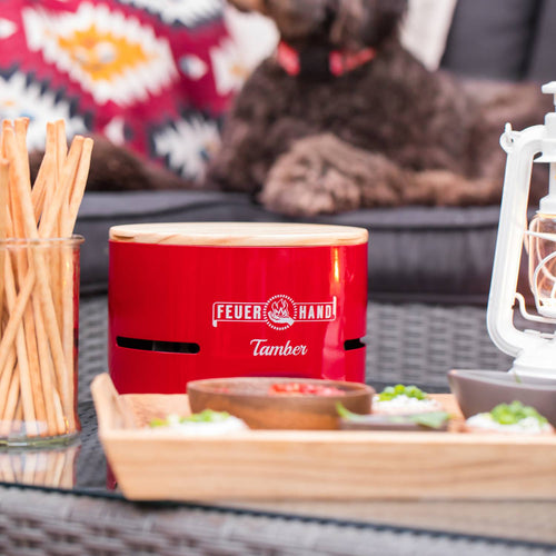 Handcrafted red Feuerhand Tamber tabletop charcoal grill with wooden lid on a glass outdoor table, surrounded by snacks and a white vintage lantern, perfect for Farm, Garden & Tool, Camping, and Grilling collections.